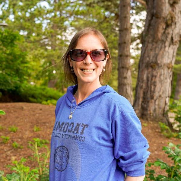 A headshot of Leigh. She smiles against a wooded background, wearing rectangular sunglasses and a royal blue "Tacoma Community College" sweatshirt.